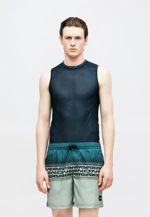 Joven con el cabello peinado hacia atrás que lleva una camiseta sin mangas de malla oscura y pantalones cortos de baño con degradado en tonos turquesa con un patrón de texto negro.