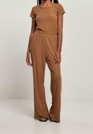 Femme portant un haut marron à manches courtes côtelées et un pantalon large assorti, debout devant un fond clair uni.