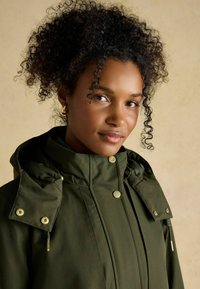 Olivewózowa wodoodporna kurtka z wysokim kołnierzem, regulowanym kapturem i złotymi napami. Charakteryzuje się gładką fakturą i dopasowanym krojem.