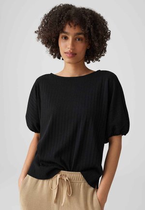 Junge Person mit lockigem Haar, die ein schwarzes Kurzarmoberteil und beige Schnürhosen trägt und vor einem schlichten grauen Hintergrund steht.