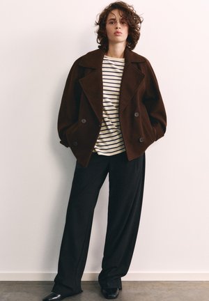 Jeune personne aux cheveux bouclés portant une veste marron oversize, un pantalon noir à jambes larges et une chemise blanche à rayures horizontales noires.