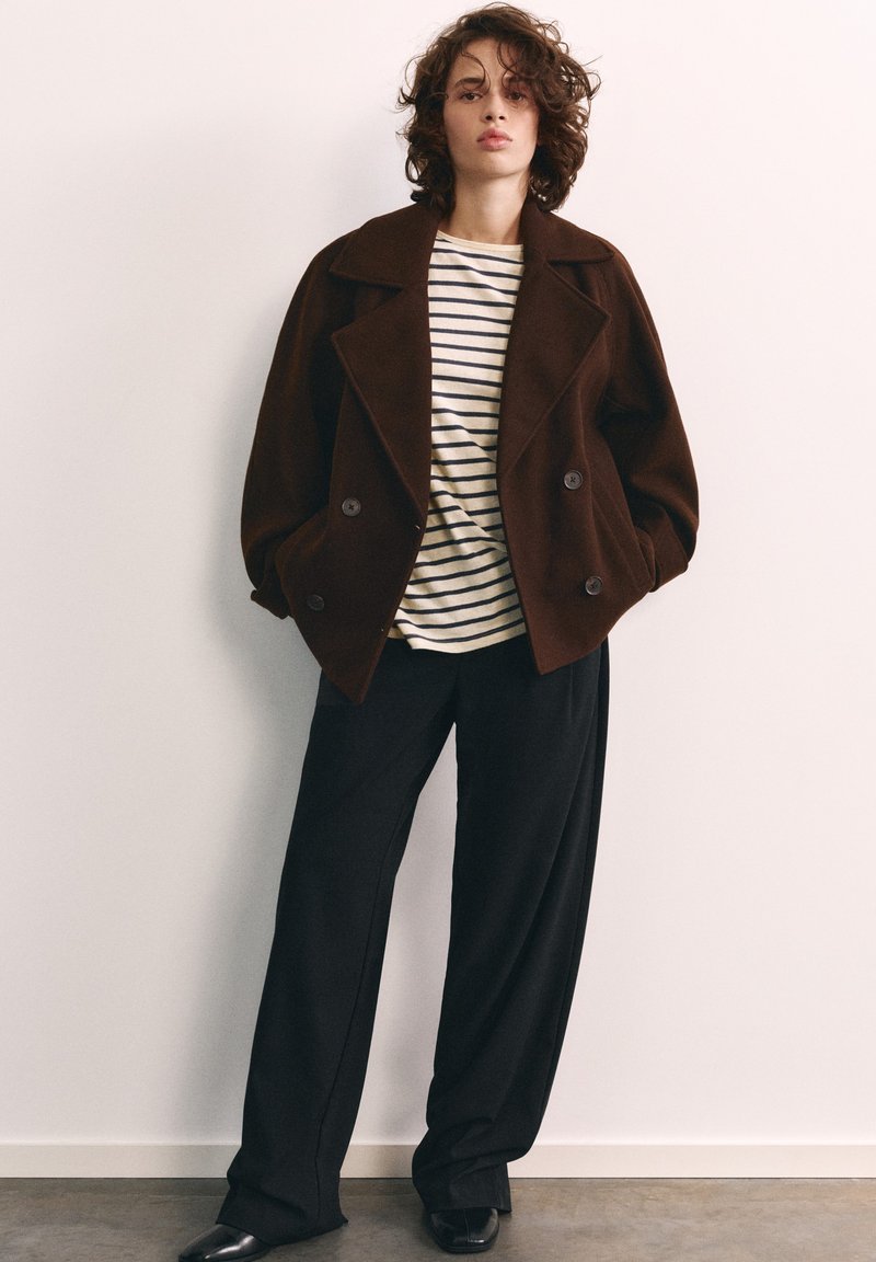 Jeune personne aux cheveux bouclés portant une veste marron oversize, un pantalon noir à jambes larges et une chemise blanche à rayures horizontales noires.
