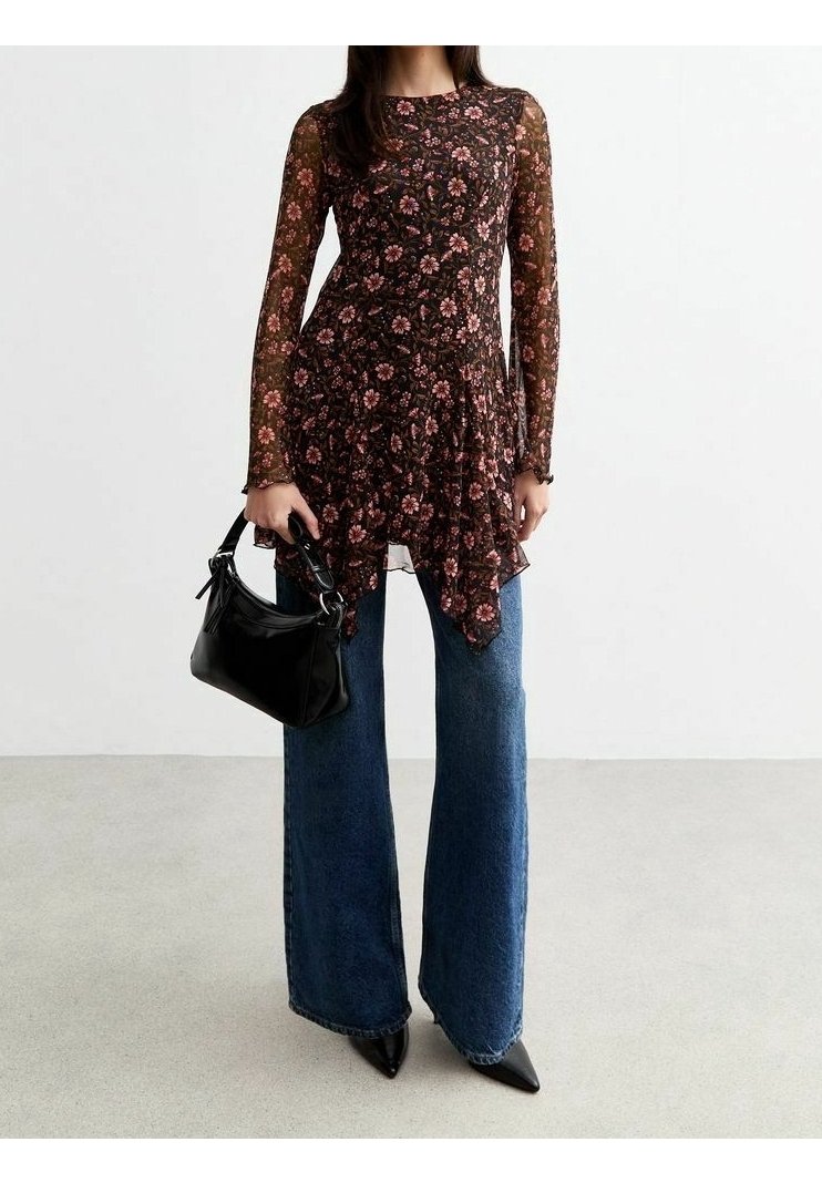 Femme portant une tunique longue à manches longues en tissu transparent à motif floral, un jean bleu à jambes larges, des chaussures noires pointues et tenant un sac à main noir.
