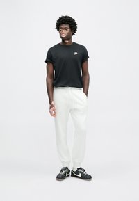 Juoda Nike marškinėliai, susukti rankogaliai, balti sportinės kelnės, juodi sportbačiai su baltais akcentais. Modelis stovi prie šviesiai pilko fono.