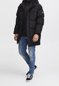 Mand går iført en sort dunjakke, mørk sweater, blå jeans og sorte-og-hvide sneakers på en simpel hvid baggrund.