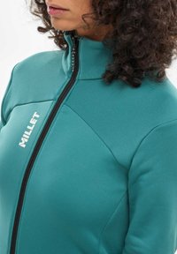Türkisfarbige Zip-Jacke aus weichem Material, mit hohem Kragen, schwarzem Reißverschluss und weißem "Millet"-Logo auf der Brust. Glatte Textur.