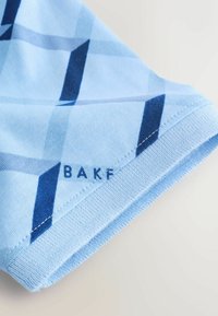 Coin en tissu bleu clair avec des rayures diagonales bleu foncé et un ourlet cousu, montrant une partie du texte "BAKF" près du bord.