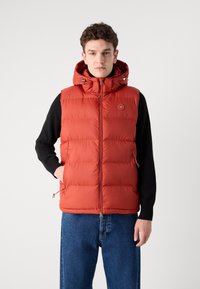 Rød puffervest med hette, glidelås foran og sidelommer. Har et quiltet design og en sirkulær logo på brystet.