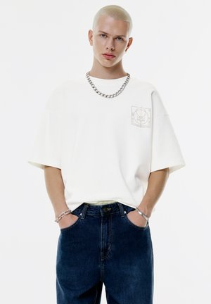 Hvid oversized t-shirt med grafik på brystet, parret med mørkeblå denim jeans og sølvfarvede kædeaccessories.