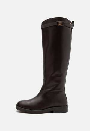 Botte haute en cuir marron foncé avec petit talon et bride décorative avec boucle métallique près du haut, présentée sur un fond uni.