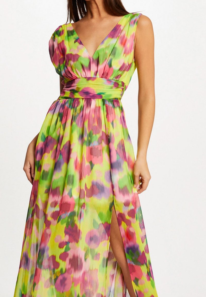 Femme portant une robe longue fleurie sans manches avec un profond décolleté en V et une fente haute sur la cuisse en vert vif, rose, violet et rouge.