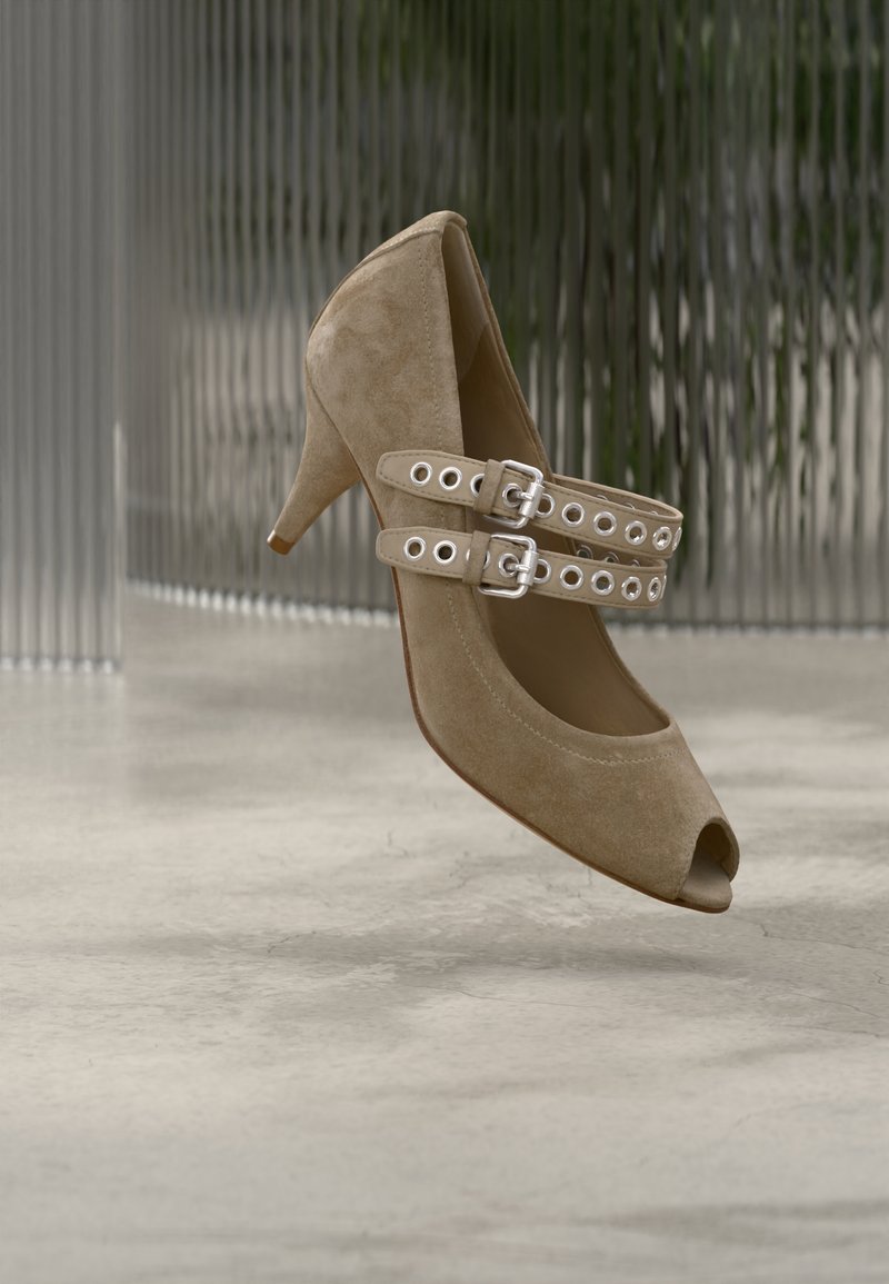 Μπεζ γόβα από σουέτ με ψηλό τακούνι και άνοιγμα στο δάχτυλο, με δύο ρυθμιζόμενους ιμάντες που έχουν κυκλικά κοψίματα και ασημένιες αγκράφες.