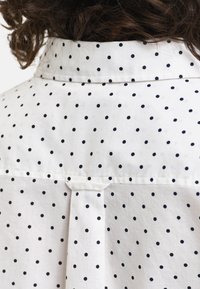 White shirt with black polka dots, featuring a buttoned collar and a pleat detail on the back, made from lightweight fabric.