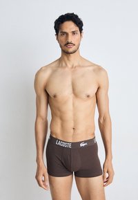 Rjavi bombažni bokserki z belim elastičnim pasom z logotipom Lacoste. Tkanina se zdi mehka in raztegljiva, tesno prilega telesu.
