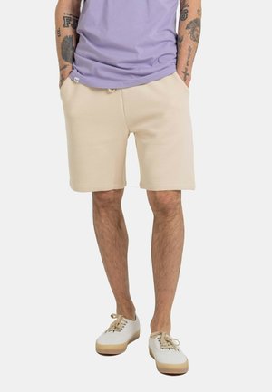 Man wearing beige textured shorts, a lavender t-shirt, white sneakers, standing with hands in pockets against a plain background.