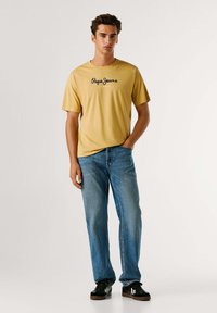 Camiseta amarilla con un logo negro de "Pepe Jeans", combinada con jeans azul claro y zapatillas negras. El diseño es casual y de corte relajado.