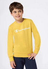 Żółta bluza z długim rękawem i białym logo "Champion", z okrągłym dekoltem, ściągaczami na rękawach i u dołu; w połączeniu z granatowymi spodniami.