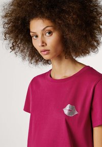 Fuksijų spalvos medvilnės marškinėliai su spausdintu lūpų dizainu metaliniu rašalu kairėje krūtinės pusėje, su apvaliu iškirpimu ir trumpomis rankovėmis.