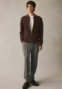 Brauner Reißverschluss-Cardigan über einem weißen Hemd, kombiniert mit grauen Streifen-Hosen und dunklen Loafers. Neutraler Hintergrund mit weicher Textur.