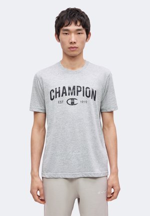 Młody mężczyzna ubrany w jasnoszary T-shirt Champion z czarnym napisem logo i pasujące jasnoszare spodnie na jednolitym tle.