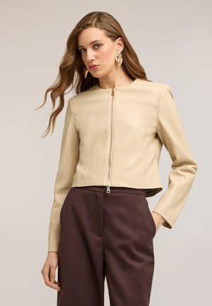 Kvinde med langt, bølget hår iført beige lynlåstrøje, mørkebrune bukser med høj talje og store guldfarvede øreringe formet som blade, poserer med den ene hånd i lommen.