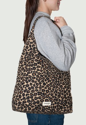 Sac fourre-tout imprimé léopard en tissu doux, aux couleurs marron et noir, avec une large bandoulière et une étiquette de marque visible cousue en bas.