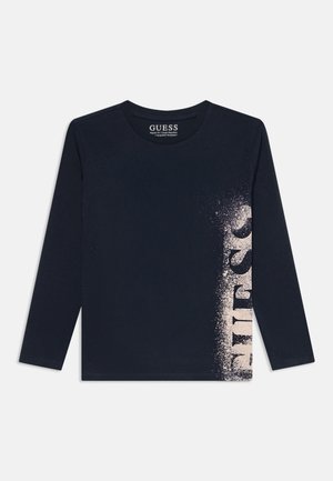 Tmavomodrá košeľa s dlhým rukávom a okrúhlym výstrihom s bielym postriekaným vertikálnym logom "GUESS" na pravej strane.