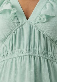 Müntroheline tekstureeritud kleit, mis sisaldab volangid, sügavat V-kaelust ja kokku pandud vööd, et luua voolav siluett.