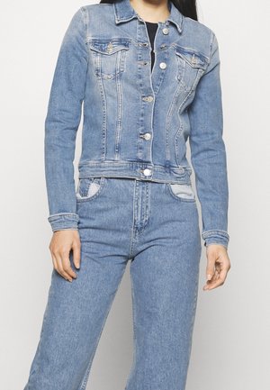 Femme portant une veste en jean bleu clair assortie et un jean, debout devant un fond clair uni.