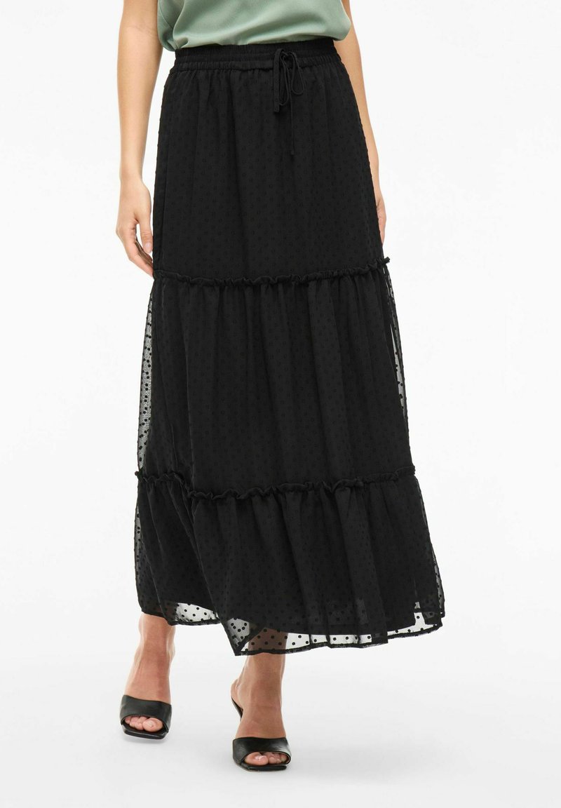 Jupe maxi noire à couches avec une texture à pois, taille élastique et bords volants. Portée avec des talons ouverts noirs.