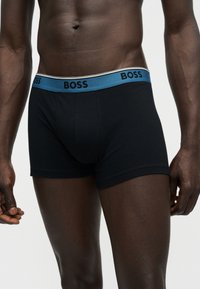 Torse d'homme à la peau foncée portant un boxer noir avec une ceinture bleue marquée "BOSS", sur un fond clair uni.