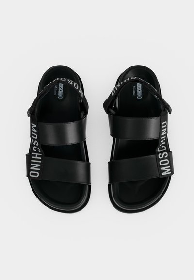Ζευγάρι μαύρα σανδάλια Moschino με χοντρές λουριά και κλεισίματα από υφασμάτινο λογότυπο σε λευκό φόντο.