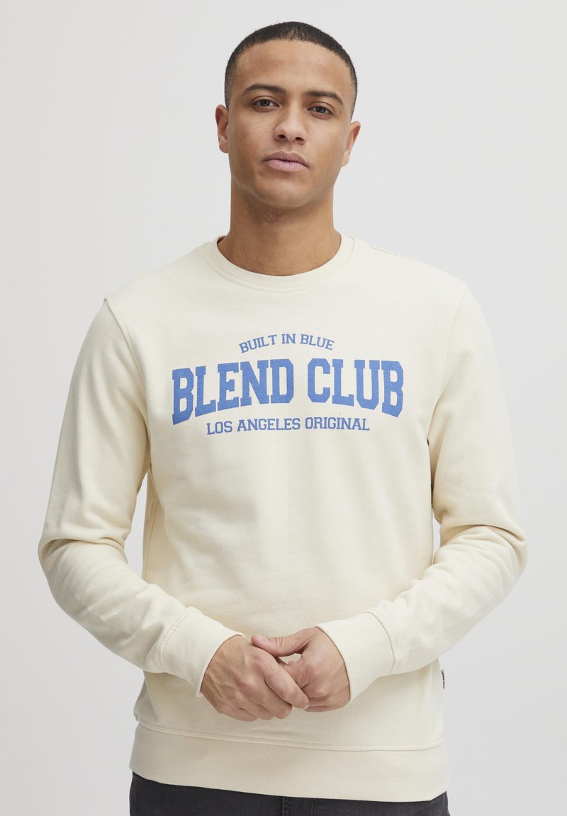 Kremfarget sweatshirt med teksten "BLEND CLUB" i blå bokstaver, rund hals og lange ermer, laget av mykt, strukturert materiale.