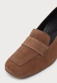 Mocassin en daim marron à bout carré arrondi, large détail de sangle sur le devant et doublure intérieure noire, présenté sur un fond blanc uni.
