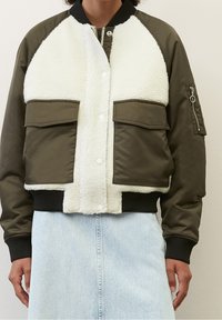 Olivgrüne Bomberjacke mit weißem Kunst-Lammfell auf der Vorderseite, gerippten Bündchen und zwei Brusttaschen. Verfügt über eine Reißverschlusstasche am linken Ärmel.