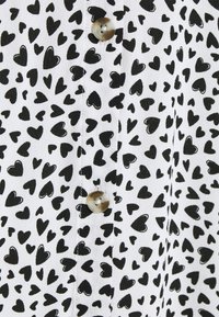 Tela blanca con un patrón de corazones negros, que presenta pequeños corazones de diferentes tamaños. Un botón de color marrón claro es visible en una tapeta.