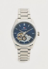 Ασημένιο μεταλλικό ρολόι χειρός με μπλε καντράν, ορατά γρανάζια, ασημένιες ενδείξεις ωρών και το λογότυπο "Hilfiger" κάτω από τη 12η ώρα, αυτόματο μηχανισμό που υποδεικνύεται στο καντράν.