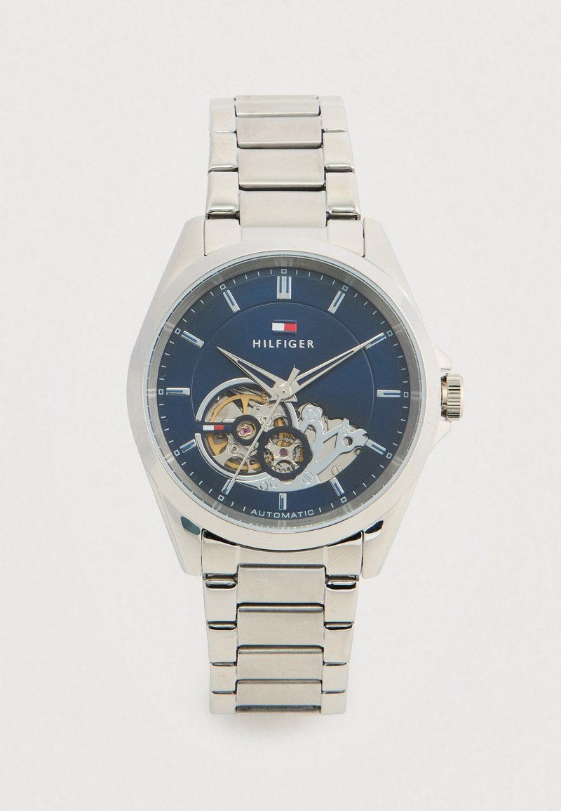 Ceas de mână din metal argintiu cu cadran albastru, cu mecanisme vizibile, markeri orari argintii și logo-ul "Hilfiger" sub 12 ore, mișcare automată indicată pe cadran.
