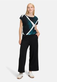 Kortärmad topp i svart med ett abstract mönster i teal och vitt, i kombination med svarta vida byxor och vita sneakers.