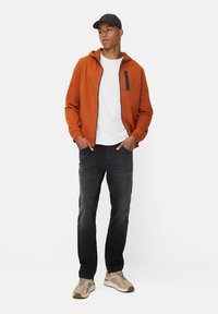 Orange fleecejakke med lynlåslomme på brystet, hvid t-shirt, sorte jeans og beige sneakers. Model iført en sort hat.