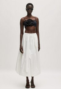 Zwarte cropped top met gerimpeld bloemen detail, gecombineerd met een witte, enkellange, verzamelde rok. Model draagt zwarte zonnebril en sandalen.