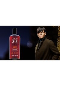 Läpinäkyvä pullo American Crew Daily Cleansing Shampoo -shampoota, 250 mL. Tumma punainen neste, musta korkki, oranssi teksti. Malli tummassa takissa pullon vieressä.
