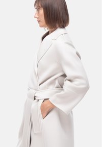 Cappotto lungo e grigio chiaro realizzato in un tessuto morbido, con vita con cintura, colletto a scialle e tasche laterali. Texture liscia con una vestibilità sartoriale.