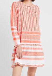 Vestido de patrones naranjas y blancos con mangas largas, que presenta diseños geométricos y rayas horizontales. Hecho de una tela ligera.