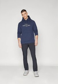 Sweat à capuche bleu marine avec poche avant et texte blanc "I can Performance", associé à un pantalon sombre et des baskets gris clair. Design simple.