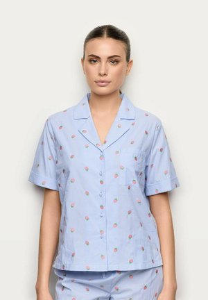 Kobieta w jasnoniebieskim, pasiastym komplecie piżamowym z krótkim rękawem i wzorem różowych róż, stojąca na jednolitym jasnym tle.
