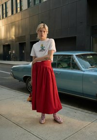 Hvit t-skjorte med blomsterbroderi, kombinert med et flytende rødt skjørt. Myke rosa snøresko og en liten veske fullfører antrekket.