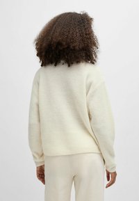 Personne avec des cheveux bouclés portant un pull en tricot côtelé crème et un pantalon crème assorti, tourné de dos contre un fond uni.