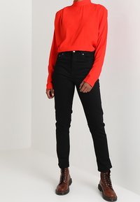 Chemise rouge à manches longues avec un col montant, associée à un pantalon noir ajusté et des bottes marron à lacets. Tissu lisse et design ajusté.