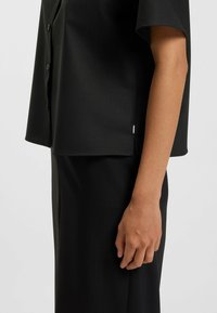 Black short-sleeve button-up shirt made of smooth fabric, featuring a relaxed fit and subtle side slit detail at the hem.